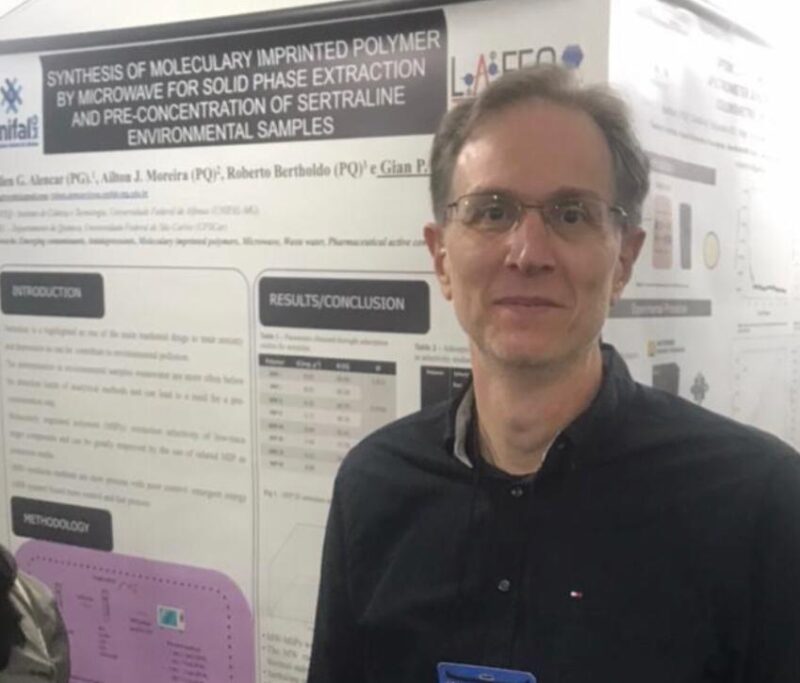 Um homem está posicionado à direita da foto, em pé, diante de um painel de apresentação científica. Ele usa óculos e camisa preta, e olha para a câmera com uma expressão neutra e leve sorriso. Ao fundo, o pôster exibe textos, gráficos e tabelas sobre uma pesquisa na área de química ou ciências ambientais, com logotipos institucionais no topo. A iluminação é suave e o ambiente parece ser um evento acadêmico ou congresso científico.