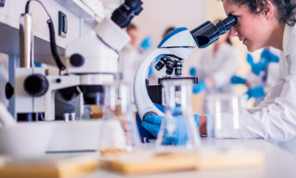 Pesquisadora em um laboratório observa uma amostra ao microscópio. Ela usa jaleco e luvas azuis. Na bancada há frascos e materiais de pesquisa, enquanto outras pessoas trabalham ao fundo, em um ambiente científico organizado.