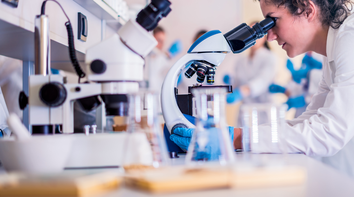 Pesquisadora em um laboratório observa uma amostra ao microscópio. Ela usa jaleco e luvas azuis. Na bancada há frascos e materiais de pesquisa, enquanto outras pessoas trabalham ao fundo, em um ambiente científico organizado.