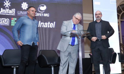Na foto, três homens participam da assinatura do Protocolo de Intenções durante o evento de formalização da Rede Sul Mineira de Ciência, Tecnologia e Inovação (RESMI), em Poços de Caldas. Ao centro, um homem de terno cinza assina o documento sobre uma mesa transparente. À esquerda, outro homem, vestido com camisa jeans azul, observa a cena. À direita, um terceiro homem, usando blazer e camisa preta, acompanha o momento. Ao fundo, um painel exibe os logotipos das instituições participantes, entre elas UNIFAL-MG, UNIFEI, CEFET-MG, UFLA, IFSULDEMINAS e Inatel.