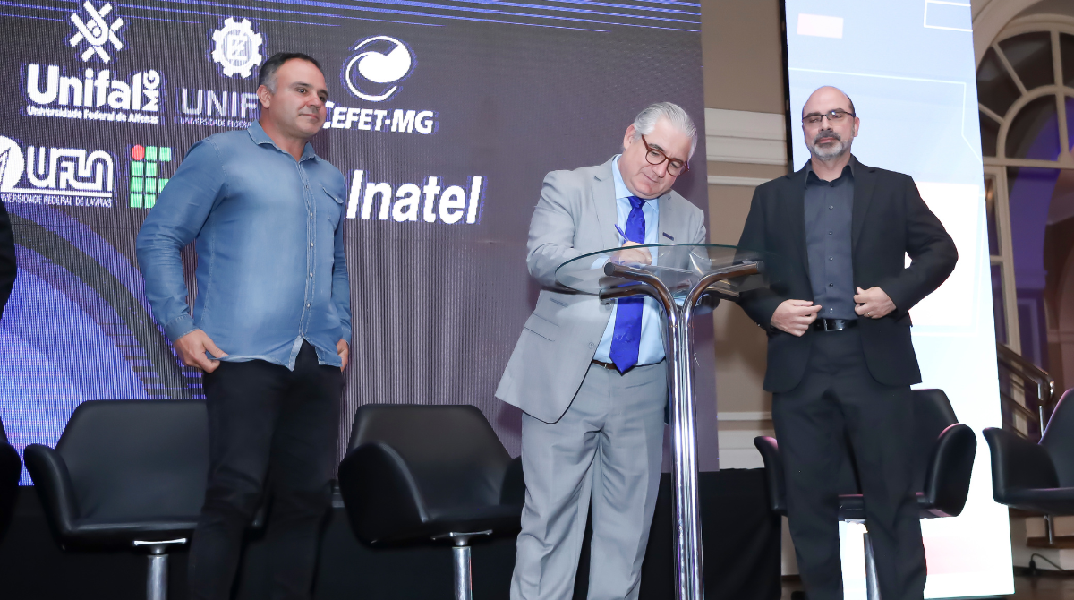 Na foto, três homens participam da assinatura do Protocolo de Intenções durante o evento de formalização da Rede Sul Mineira de Ciência, Tecnologia e Inovação (RESMI), em Poços de Caldas. Ao centro, um homem de terno cinza assina o documento sobre uma mesa transparente. À esquerda, outro homem, vestido com camisa jeans azul, observa a cena. À direita, um terceiro homem, usando blazer e camisa preta, acompanha o momento. Ao fundo, um painel exibe os logotipos das instituições participantes, entre elas UNIFAL-MG, UNIFEI, CEFET-MG, UFLA, IFSULDEMINAS e Inatel.
