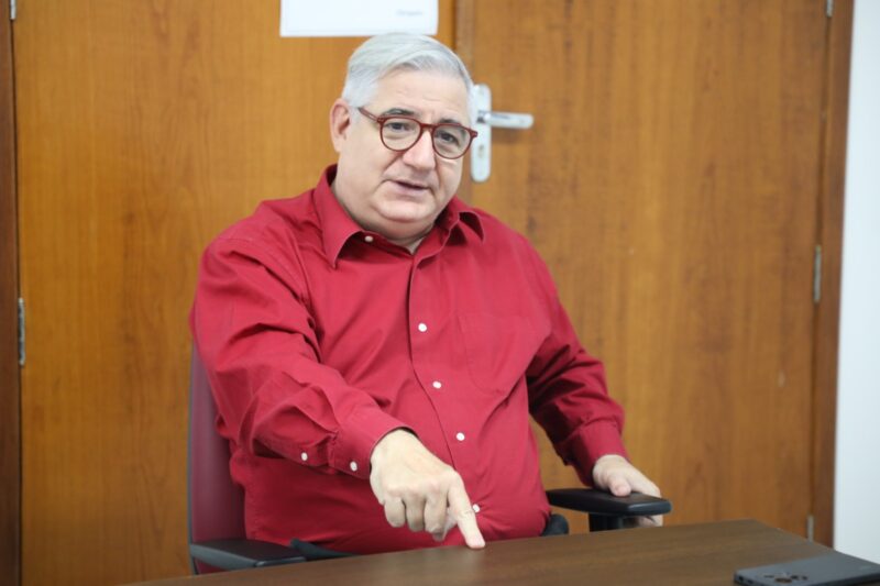 A foto mostra um homem sentado, Professor Sandro Cerveira, em uma sala, diante de uma mesa. Ele tem cabelos curtos grisalhos e usa óculos de armação vermelha. Veste uma camisa social vermelha de mangas compridas. Na imagem, ele gesticula com a mão direita apontando para a mesa, como se explicasse ou enfatizasse algum ponto durante uma conversa. Ao fundo, há uma porta de madeira clara com uma placa branca na parte superior.
