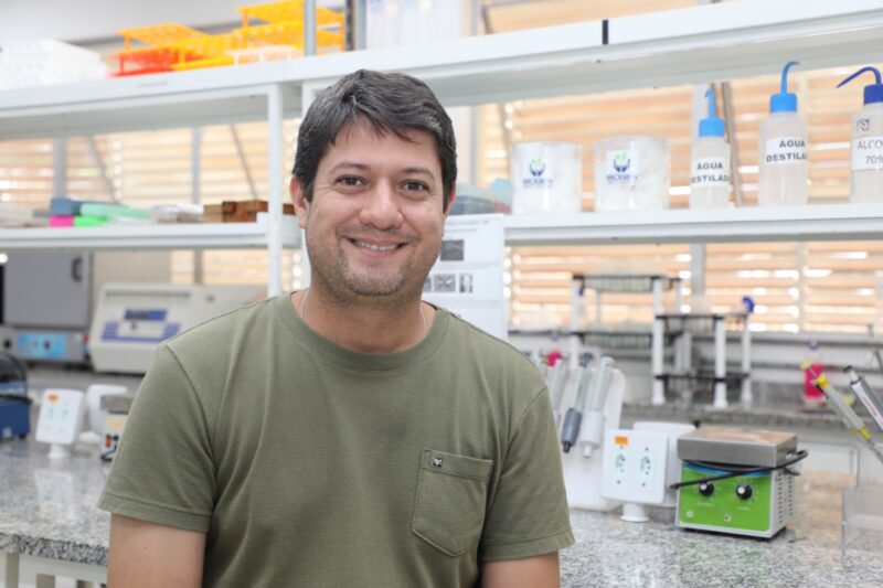 O professor Thiago Corrêa está sentado em um laboratório, sorrindo para a câmera. Ele veste uma camiseta verde e aparece em primeiro plano. Ao fundo, é possível ver bancadas com equipamentos de pesquisa, frascos rotulados e materiais laboratoriais organizados nas prateleiras, compondo um ambiente científico claro e bem iluminado.