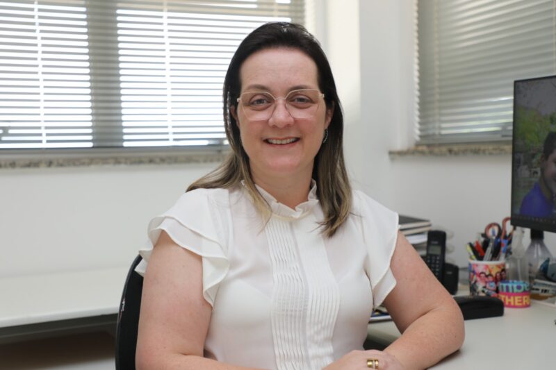 A foto apresenta uma mulher está sentada em um escritório iluminado, sorrindo para a câmera. Ela usa uma blusa branca de mangas curtas com detalhes plissados na parte frontal e óculos de armação clara. Ao fundo, há janelas com persianas parcialmente fechadas, uma mesa com computador, porta-canetas e alguns documentos organizados. O ambiente transmite sensação de organização e profissionalismo.