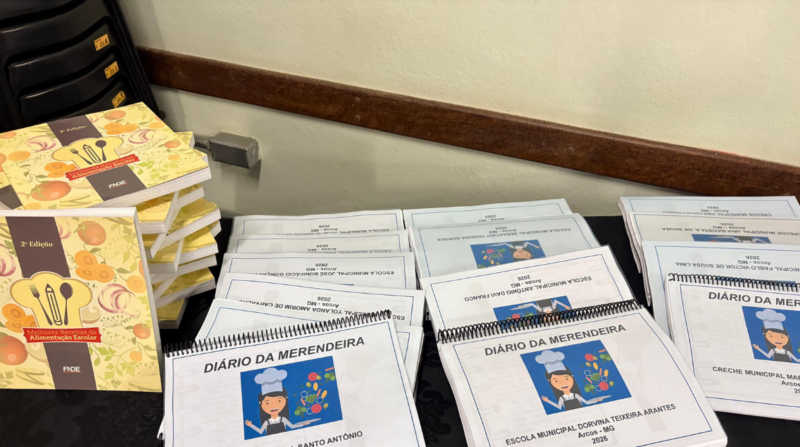 Mesa com materiais distribuídos para capacitação, incluindo exemplares do livro “Melhores Receitas da Alimentação Escolar” e vários cadernos encadernados intitulados “Diário da Merendeira”, identificados com nomes de escolas municipais de Arcos – MG e ano de 2026.