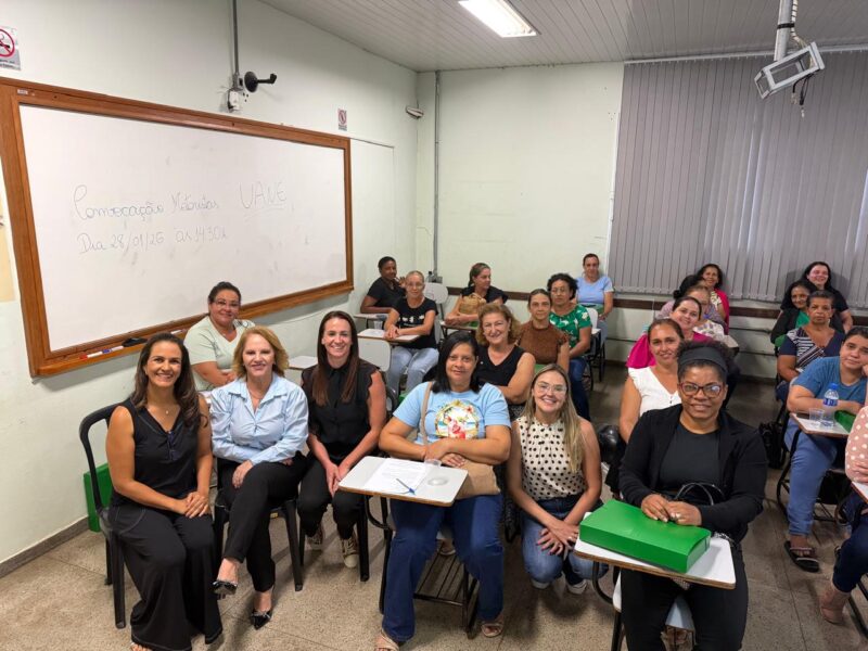 Grupo de merendeiras reunidas em sala de aula durante capacitação da rede municipal. As participantes estão sentadas em carteiras com caixas verdes e sorriem para a foto, junto ao instrutor posicionado na frente da turma.