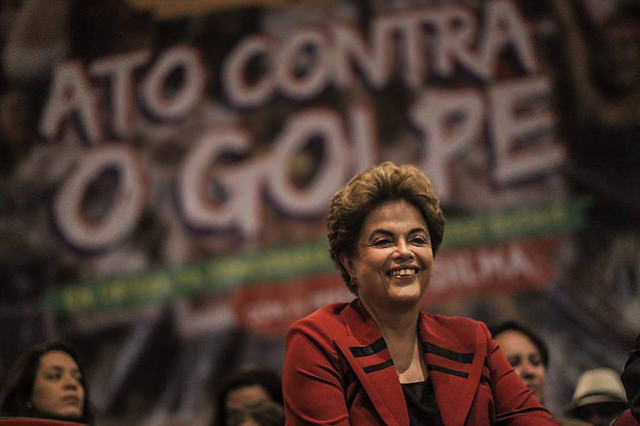 Fotografia colorida de um evento político. Em primeiro plano, uma mulher, Dilma Roussef, cabelos castanhos curtos e volumosos, sorri enquanto está sentada. Ela veste um blazer vermelho com detalhes pretos sobre uma blusa escura. Sua expressão é serena e confiante.Ao fundo, levemente desfocado, há um grande painel com a frase “ATO CONTRA O GOLPE” escrita em letras brancas de destaque. Outras pessoas aparecem sentadas atrás dela, mas não estão em foco. A iluminação concentra-se na mulher, destacando seu rosto e sua roupa em contraste com o fundo mais escuro.