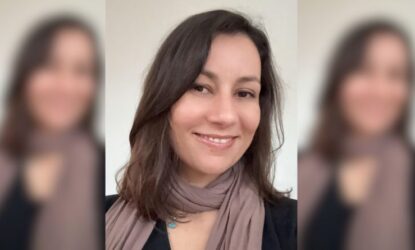Uma mulher jovem adulta, de pele clara e cabelo castanho escuro na altura dos ombros, levemente ondulado e repartido de lado, sorri suavemente para a câmera. Ela veste uma blusa escura e um lenço marrom claro enrolado no pescoço. O fundo é neutro e claro, com as laterais da imagem levemente desfocadas.
