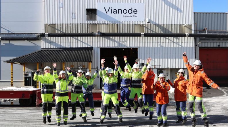 Um grupo de trabalhadores da indústria posa em frente a um prédio industrial com a placa “Vianode Industrial Pilot”. Eles usam capacetes brancos e roupas de segurança em cores chamativas, como verde-limão e laranja, com faixas refletivas. Todos aparecem pulando com os braços levantados, demonstrando entusiasmo. Ao fundo, há a fachada metálica do galpão, uma porta aberta e parte de um equipamento ou veículo industrial à esquerda.