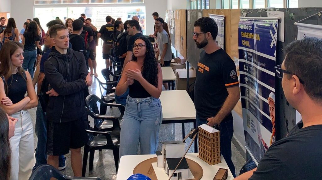 Grupo de estudantes reunidos em um espaço interno, observando a apresentação de um projeto. No centro, uma jovem de óculos e blusa preta gesticula enquanto explica algo. Ao lado dela, um homem com barba e camiseta preta segura uma maquete de edifício. Sobre a mesa há uma maquete arquitetônica com prédios em miniatura. Ao fundo, há cartazes e painéis informativos, incluindo um banner sobre curso de Engenharia Civil, e várias outras pessoas conversando.