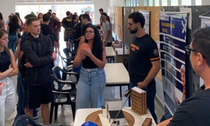 Grupo de estudantes reunidos em um espaço interno, observando a apresentação de um projeto. No centro, uma jovem de óculos e blusa preta gesticula enquanto explica algo. Ao lado dela, um homem com barba e camiseta preta segura uma maquete de edifício. Sobre a mesa há uma maquete arquitetônica com prédios em miniatura. Ao fundo, há cartazes e painéis informativos, incluindo um banner sobre curso de Engenharia Civil, e várias outras pessoas conversando.